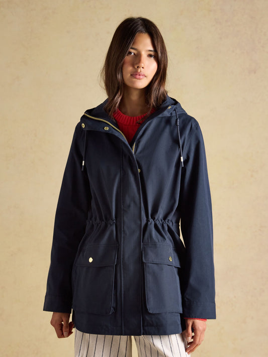 Woman wearing a navy Joules Portwell waterproof raincoat with a peaked hood and drop-tail hem on a coastal walk.