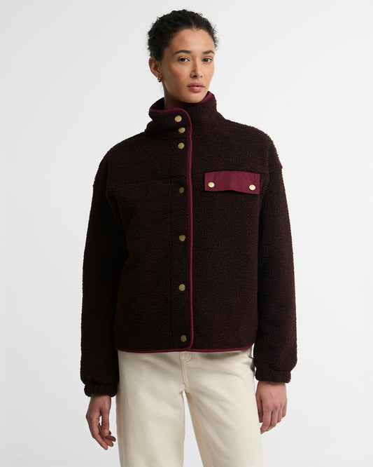 Woman wearing a deep burgundy Barbour Mabel button-up fleece bomber jacket with a high collar, contrasting red trim, and brass buttons.
