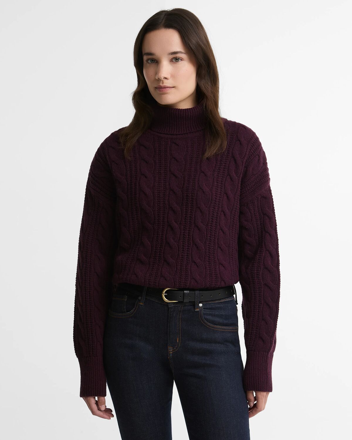 A woman wearing the burgundy Barbour Agatha Knit Jumper, featuring a textured cable knit roll neck pattern, tucked into dark denim jeans with a black belt.