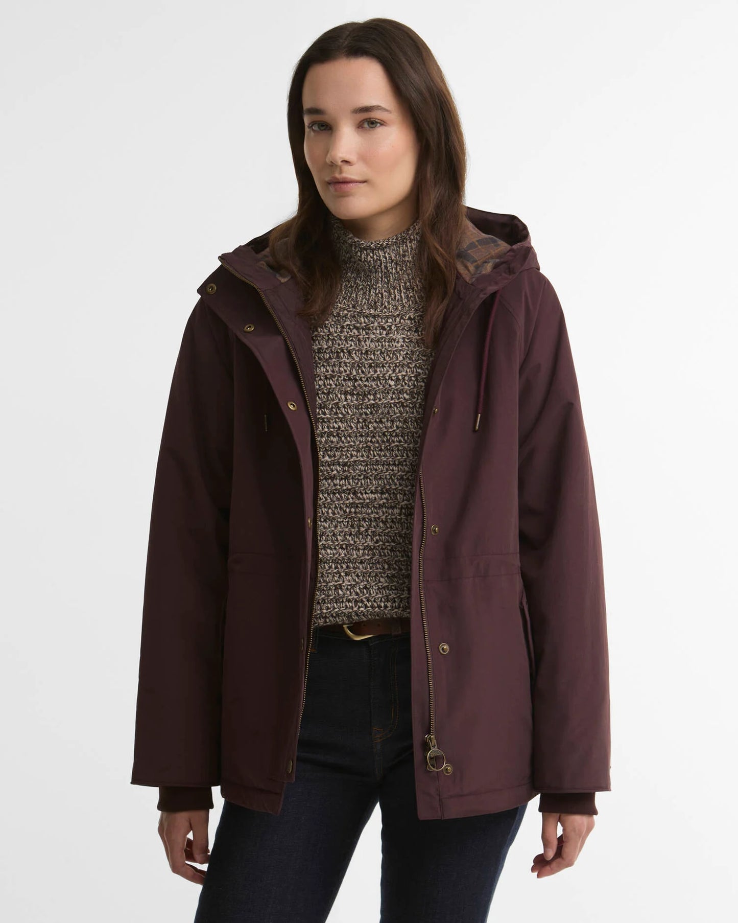 A woman wearing a dark plum hooded jacket over a textured gray knit sweater and dark jeans, photographed against a clean light gray studio background.