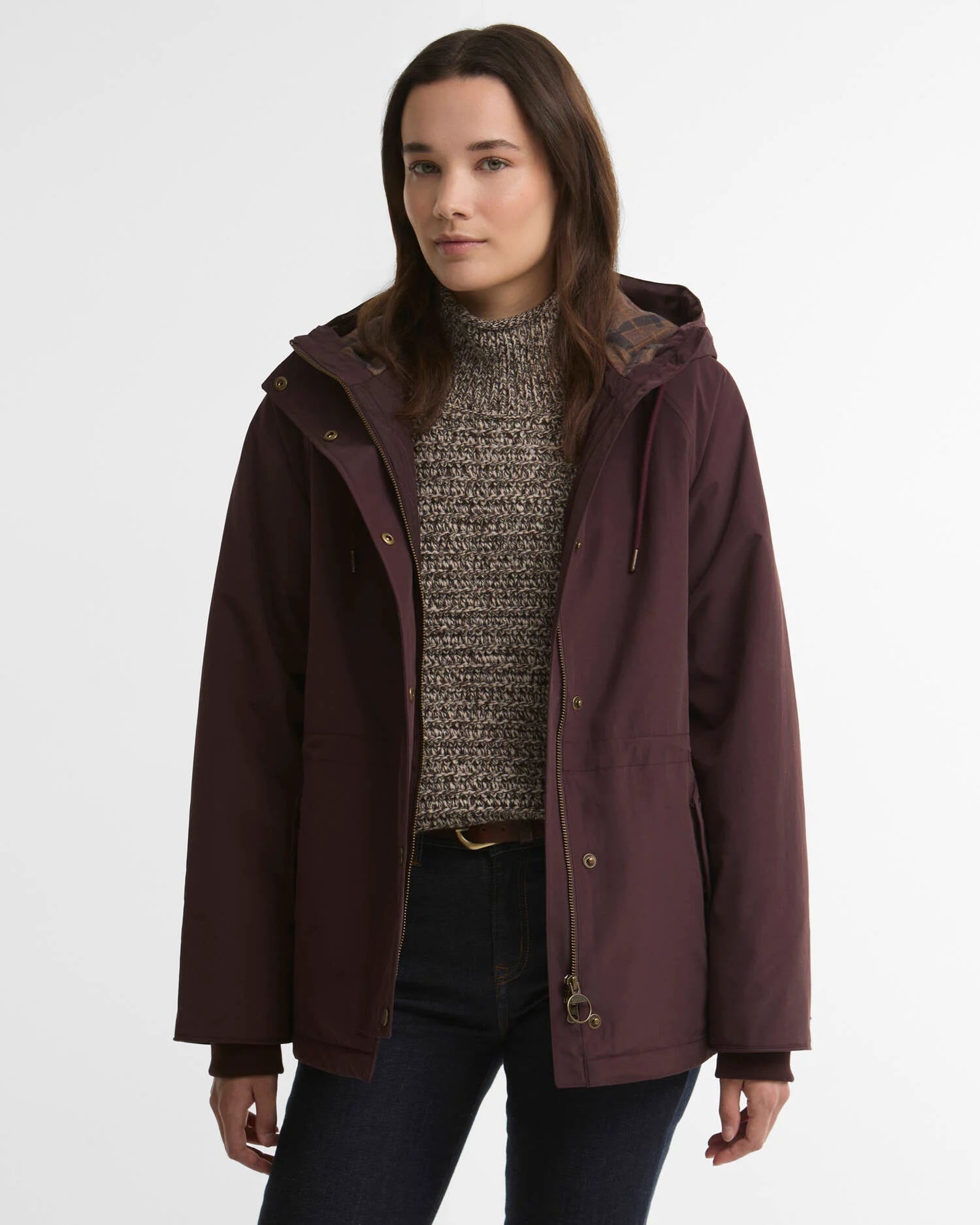 A woman wearing a dark plum hooded jacket over a textured gray knit sweater and dark jeans, photographed against a clean light gray studio background.