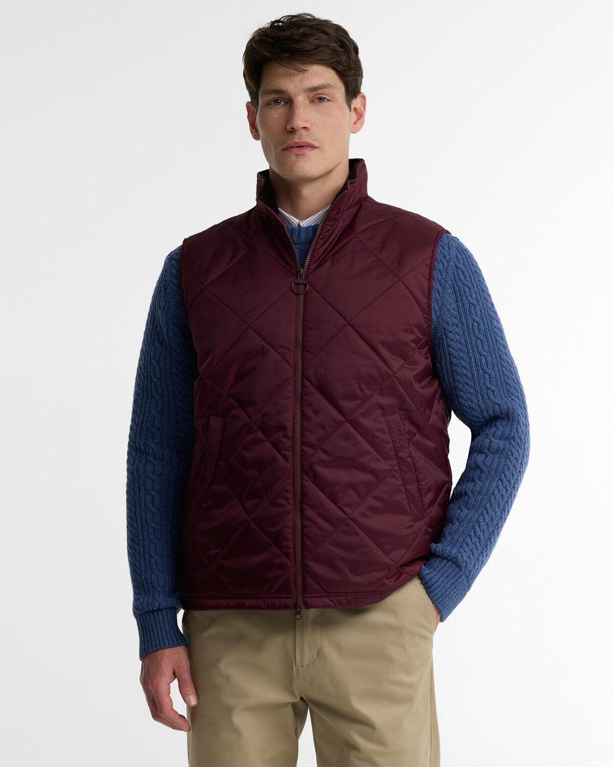 A man wearing a deep burgundy Barbour Finn box-quilted gilet over a blue cable knit sweater.