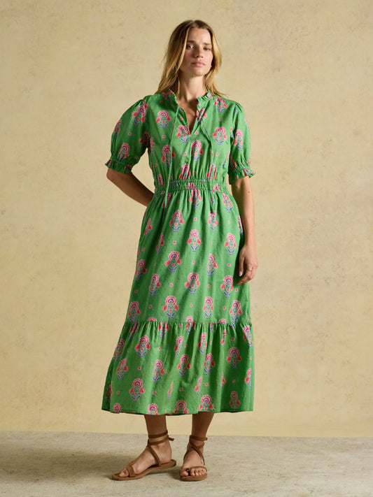Front view of a woman wearing the green Joules Larissa Tie Neck Midi Dress with a pink floral pattern, paired with brown strappy sandals.
