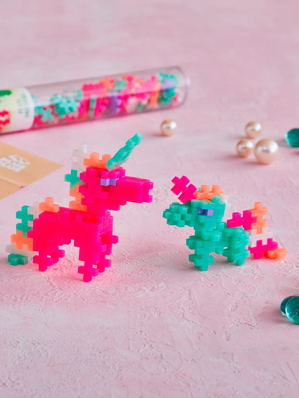 Plus-Plus Pearl Unicorns clear storage tube standing next to constructed pink and green toy unicorn figures.