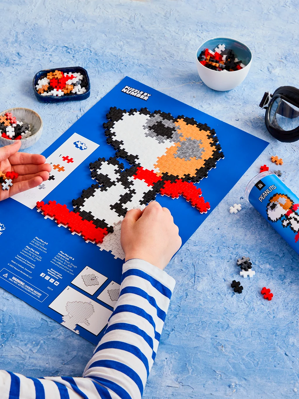 Plus-Plus Snoopy Flying Ace tube container alongside the assembled pixel-style Snoopy artwork featuring his aviator cap and red scarf.