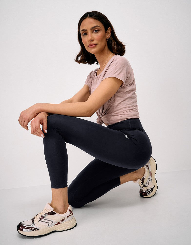 Side view of a woman crouching while wearing black Crew Clothing High Waisted 7/8 Leggings, paired with a light pink t-shirt and trainers.
