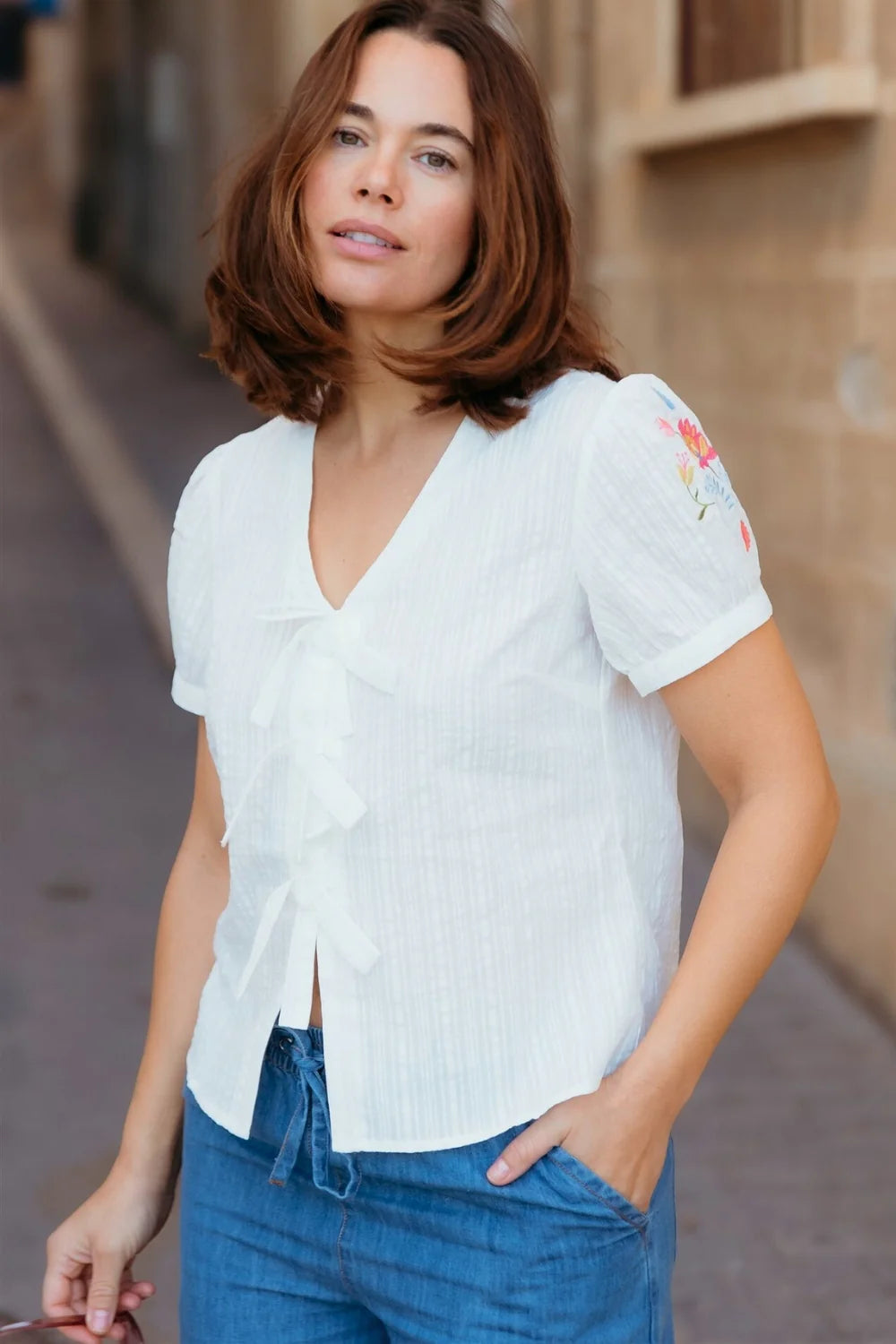 Woman wearing a white Sugarhill Brighton Adaline cotton top with embroidered floral details on the sleeve and decorative bow ties down the front.