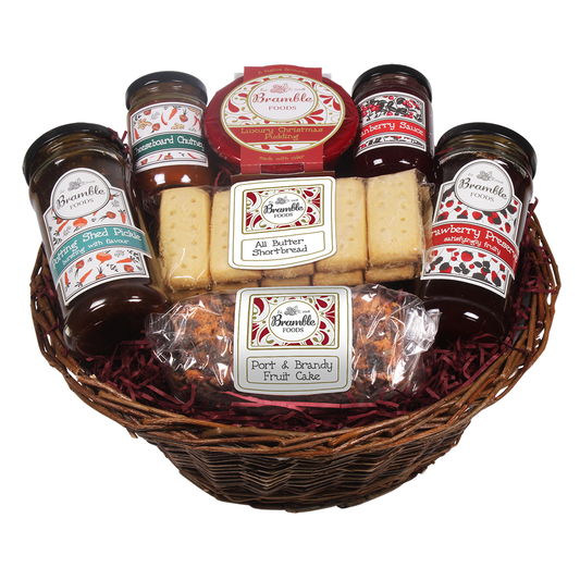 A wicker basket containing an assortment of food items including jars of pickles, chutney and cranberry sauce, shortbread fingers, a packet of port and brandy fruit cake, and a Christmas pudding.