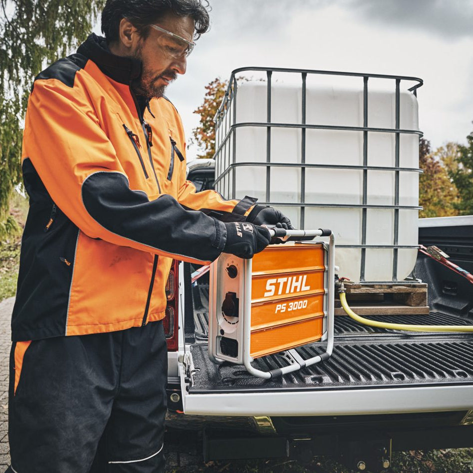 A man in an orange and black Stihl work jacket stands at the tailgate of a pickup truck outdoors, handling an orange and silver Stihl FS 360 X2 chainsaw. A large white water tank is visible in the truck bed behind him.
