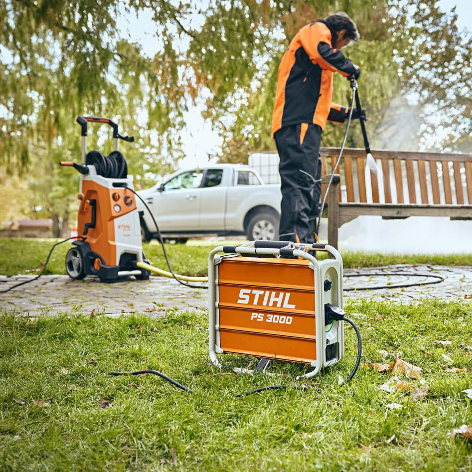 A person in an orange and black uniform operates a STIHL PS 3000 pressure washer outdoors on a grassy lawn, with another orange and white STIHL pressure washer and a white pickup truck visible in the background near a wooden bench.